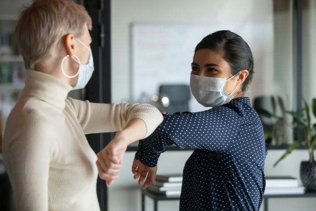 Två kvinnor med munskydd hälsar på varandra med armbågarna på ett kontor.
