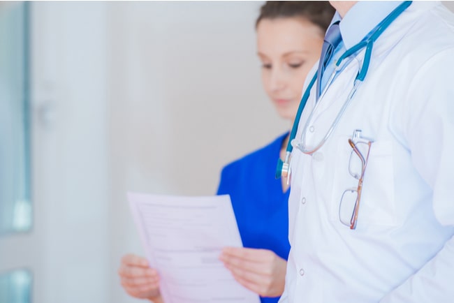 Läkare i förgrunden med en sköterska i bakgrunden som tittar på papper.