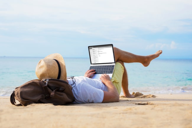 man ligger på strand med laptop i händerna.